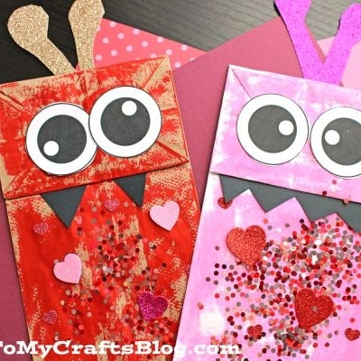 Two paper bag love monsters—one red and one pink—feature googly eyes, heart shapes, glitter, and jagged black mouths, displayed atop layered pink and red paper backgrounds.