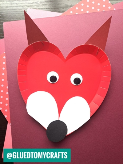 A red paper plate heart craft shaped like a fox’s face with pointy ears, big googly eyes, a white snout, and a black nose, displayed on pink and red paper backgrounds.