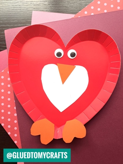 A charming paper plate heart craft: a red plate cut into a heart shape, decorated with googly eyes, an orange beak, white belly, and orange feet to create a cute bird, set on pink and purple paper.