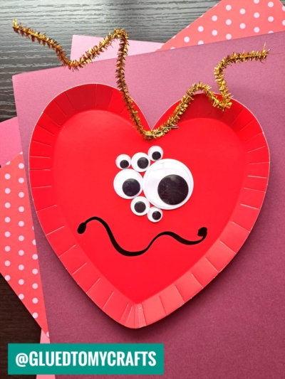 A red, heart-shaped paper plate is decorated with various sizes of googly eyes, a black squiggly smile, and a gold twisted pipe cleaner as an antenna—an adorable idea for paper plate heart crafts, resting on pink and red papers.