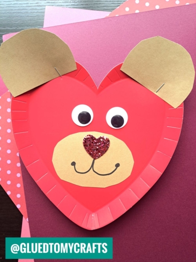 A heart-shaped red paper plate heart craft decorated as a bear with brown paper ears, googly eyes, a tan snout, a glittery heart-shaped nose, and a drawn mouth, placed on pink and patterned paper sheets.