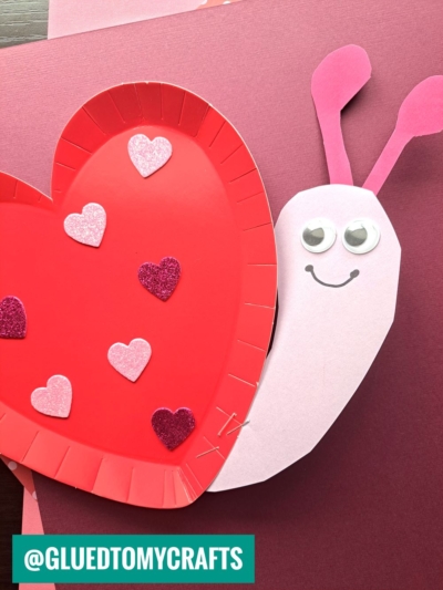 A snail craft made from a red paper plate and pink paper, decorated with googly eyes and glittery heart stickers, is placed on maroon sheets. Perfect for paper plate heart crafts! The Instagram handle @GLUEDTOMYCRAFTS is visible at the bottom.