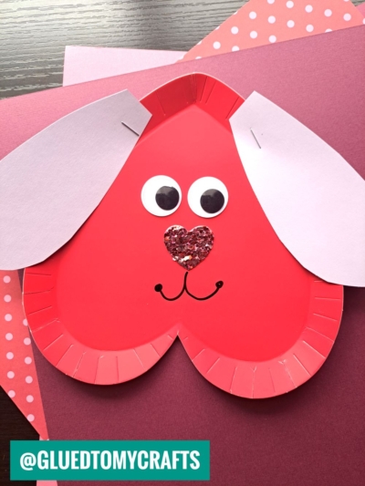 This paper plate heart craft features a red heart-shaped plate transformed into a dog with white paper ears, googly eyes, a glittery heart nose, and a drawn smile, displayed on layered pink and red sheets.