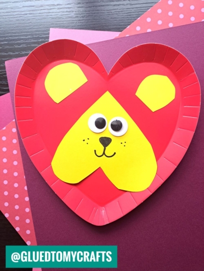 This paper plate heart craft features a red, heart-shaped plate decorated with yellow paper shapes for a lion’s face, complete with googly eyes and a drawn nose and mouth, resting on pink and purple polka dot papers.