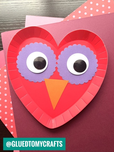 A red, heart-shaped paper plate owl craft features purple scalloped circles and large googly eyes for the face, plus an orange triangle beak. Perfect for paper plate heart crafts, with pink patterned paper as the backdrop.