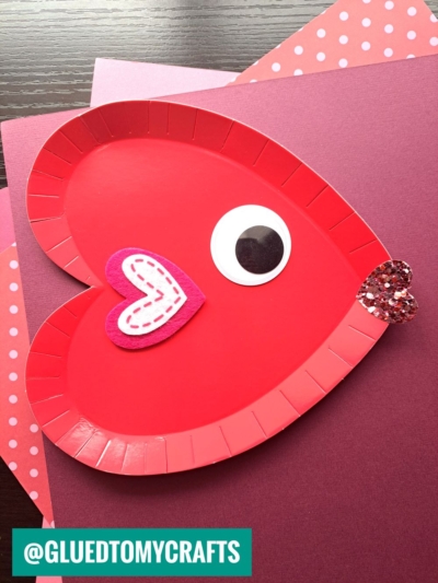 A red paper plate heart craft cut and decorated to look like a heart-shaped fish, featuring a large eye, pink felt gill, and glittery mouth, placed on pink and purple paper sheets.