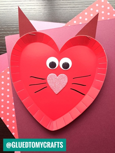 A red paper plate heart is decorated as a cat with pointy ears, googly eyes, black whiskers, and a pink glittery heart nose—perfect inspiration for fun paper plate heart crafts on patterned and solid colored paper.