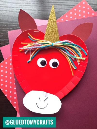 A red paper plate unicorn craft with googly eyes, a gold glitter horn, colorful yarn mane, and paper ears and muzzle, featured on layered red and pink paper—perfect for fans of paper plate heart crafts. @GLUEDTOMYCRAFTS is visible in the bottom corner.