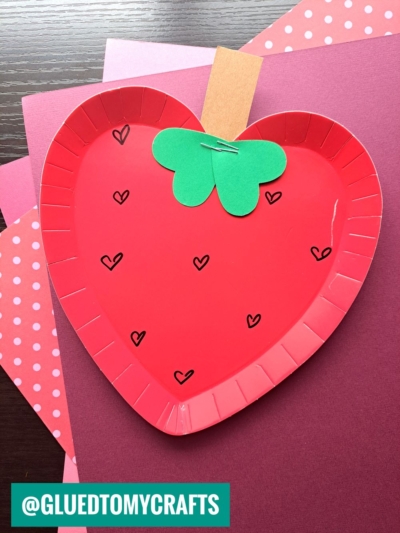 A red paper plate heart craft decorated to look like a strawberry, with small heart shapes drawn on it, a green paper leaf, and a brown paper stem, placed on pink and purple paper backgrounds.
