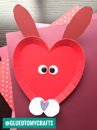 A heart-shaped red paper plate is decorated as a bunny with two large ears, googly eyes, and a white nose on purple and polka dot paper—perfect for paper plate heart crafts. @GLUEDTOMYCRAFTS is labeled in the bottom left corner.