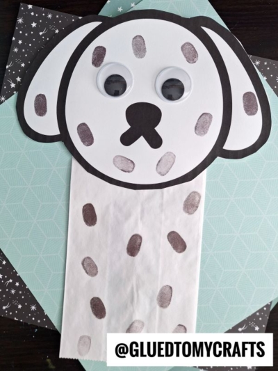A paper bag dalmatian craft with a white face, floppy black-outlined ears, large googly eyes, and marker spots, set on patterned paper backgrounds. @GLUEDTOMYCRAFTS is written at the bottom.