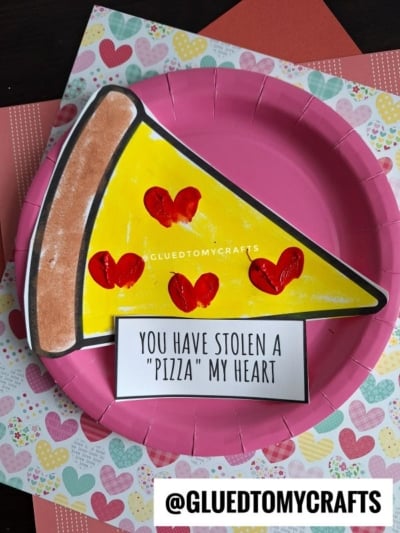 A paper craft pizza slice with heart-shaped pepperonis sits on a pink paper plate. Below it is a note that says, “You have stolen a ‘pizza’ my heart.” Craft materials with heart patterns create a fun Valentine’s Day vibe in the background.