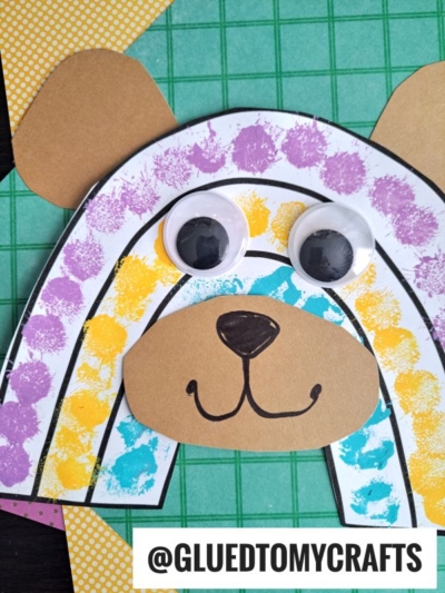 A colorful rainbow bear craft made from paper features a brown face, large googly eyes, and ears. The bear’s body has purple, yellow, and blue painted stripes on white paper. The username @GLUEDTOMYCRAFTS is at the bottom.