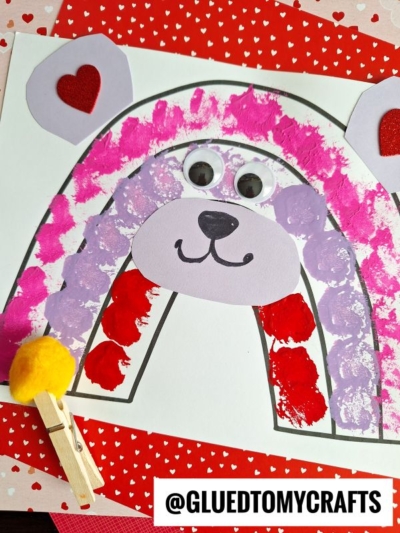 A kids’ craft of a rainbow bear made with painted dots in pink, red, and purple, large googly eyes, a purple oval snout, and red heart-shaped ears. A clothespin with a yellow pom-pom and “@GLUEDTOMYCRAFTS” are also shown.