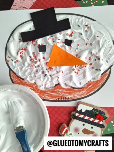 A puffy paint snowman face made with white shaving cream, a black paper hat, orange carrot nose, and red sprinkle decorations. A paintbrush and shaving cream are on the side, next to a snowman donut cookie decoration.