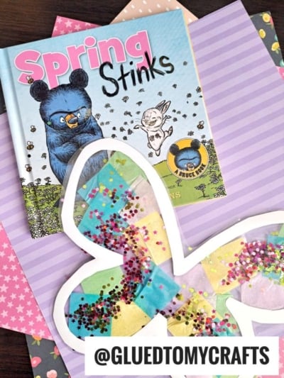 A childrens book titled Spring Stinks sits on pastel patterned paper with a butterfly suncatcher craft made of tissue paper and glitter. The handle @GLUEDTOMYCRAFTS appears at the bottom.