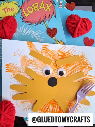 A fork painted Lorax craft with googly eyes and orange paint sits on a table with The Lorax book, red yarn hearts, glitter heart cutouts, and a purple fork. Text reads @GLUEDTOMYCRAFTS.