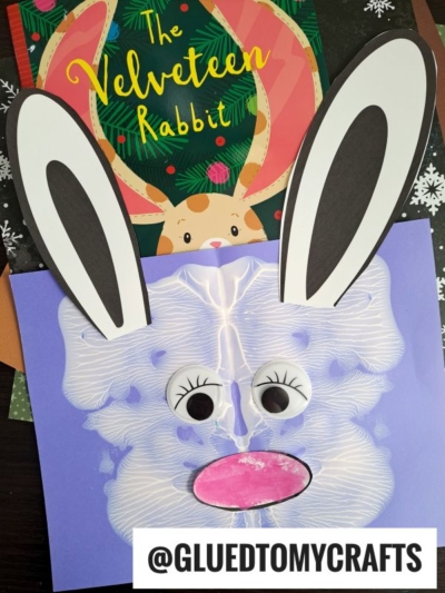 A children’s book, The Velveteen Rabbit, is displayed behind a paint splat bunny craft made from shaving cream art, large paper ears, googly eyes, and a pink nose. The username @GLUEDTOMYCRAFTS appears at the bottom.