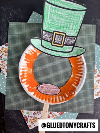 A paper plate leprechaun mask features an orange painted beard with a cut-out center and a colored paper leprechaun hat on top, all mounted on a stick. The background is decorated with patterned scrapbook paper.