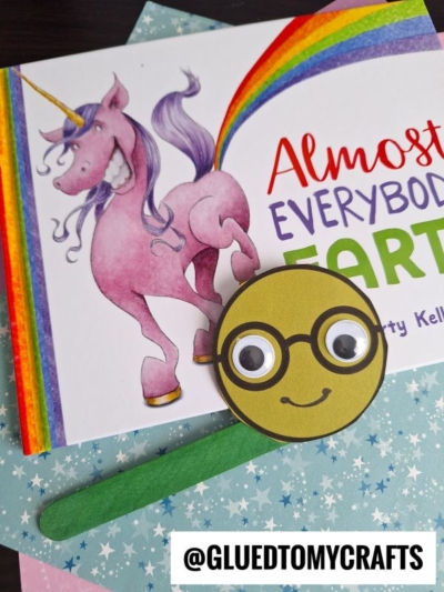 A children's book with a smiling unicorn next to a rainbow and the title Almost Everybody Farts. In front, there is a cute popsicle stick bookworm craft with a smiley face, glasses, and googly eyes on a green stick.