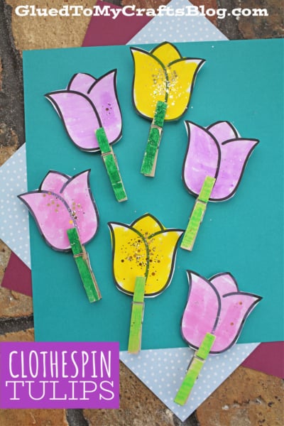 A flat lay of six Clothespin Tulip Flowers—three yellow and three purple—crafted from paper and attached to green-painted clothespins, arranged on layered sheets of colored paper. Text reads: CLOTHESPIN TULIPS.
