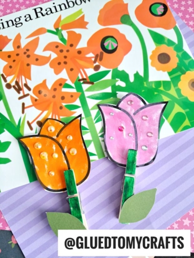 Colorful handmade flower crafts, including a Clothespin Tulip Flower, are displayed on a purple-striped surface, with an illustrated flower book page in the background. Text reads @GLUEDTOMYCRAFTS.