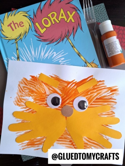A handmade fork painted Lorax craft featuring orange handprint cutouts, large googly eyes, a paper nose, and marker strokes, displayed on a copy of The Lorax book with paint, a fork, and the @GLUETOMYCRAFTS tag.