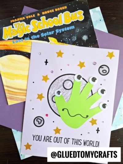 A children’s space-themed craft featuring a playful green handprint alien with googly eyes, sparkling stars, and the message “YOU ARE OUT OF THIS WORLD!” displayed beside a Magic School Bus book.