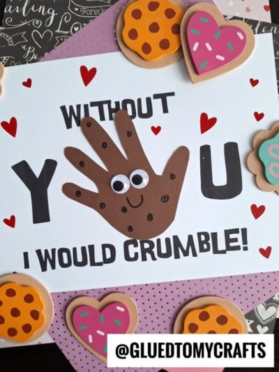 A handmade card with the message Without you I would crumble! featuring a brown handprint cookie with googly eyes and chocolate chip details as the letter “O.” Surrounded by paper hearts and cookie decorations. @GLUEDTOMYCRAFTS is at the bottom.