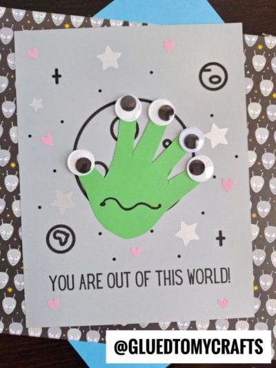 A handmade card featuring a green handprint alien, googly eyes, and a wavy mouth. The card says YOU ARE OUT OF THIS WORLD! with space-themed designs. @GLUEDTOMYCRAFTS is printed at the bottom.