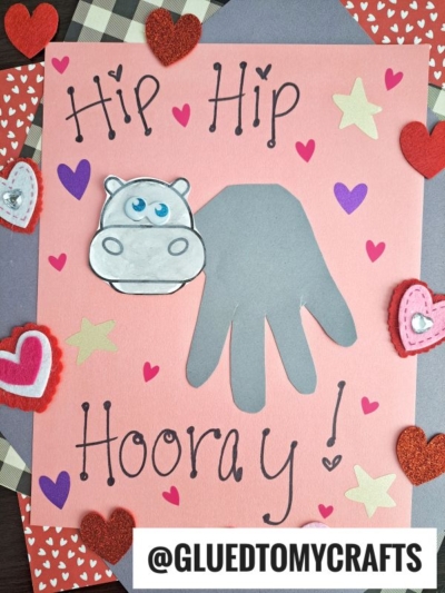 A pink card decorated with foam hearts and stars, featuring the words Hip Hip Hooray!, a gray handprint hippo design, and a cute hippo drawing. The handle @GLUEDTOMYCRAFTS is displayed at the bottom.
