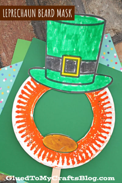 A handmade paper plate leprechaun mask with a green top hat and orange beard, crafted from paper and colored with markers, is shown resting on green and polka-dotted paper backgrounds.