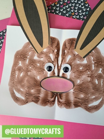 A paint splat bunny face made from brown paint blots on white paper, with large paper ears, googly eyes, and a pink nose, set atop pink and black heart-patterned papers. @GLUETOMYCRAFTS label is at the bottom left.