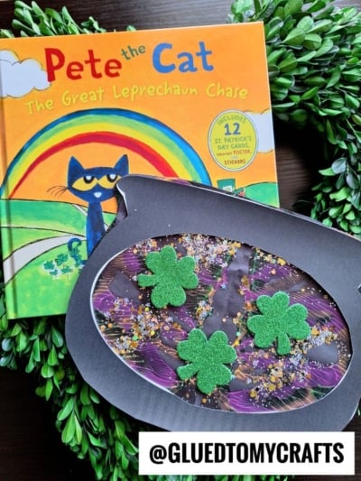 A Pete the Cat book titled The Great Leprechaun Chase rests on a green wreath next to a Paint Splat Leprechaun Cauldron craft, decorated with glitter and green foam shamrocks. The username @GLUEDTOMYCRAFTS appears at the bottom.