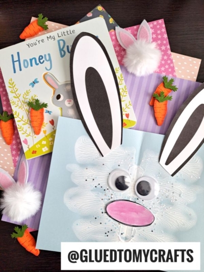 Children’s books, bunny-themed crafts like a paint splat bunny, felt carrots, and a handmade bunny card with large ears, googly eyes, and a pom-pom nose are arranged on pastel-striped paper backgrounds. @GLUEDTOMYCRAFTS is at the bottom.