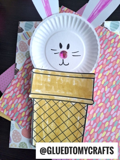 A bunny ice cream cone face made from a paper plate with drawn whiskers and nose, attached to a paper cone, rests on Easter-themed and polka dot craft paper.