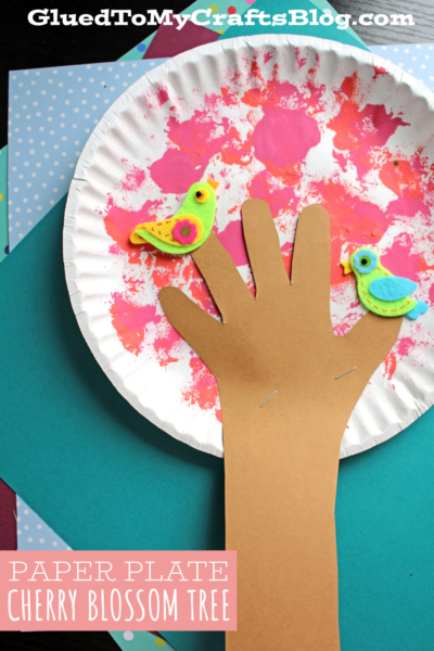 A paper plate cherry blossom tree is created by painting a plate pink and white for blossoms, then stapling on a brown paper hand as the trunk and branches. Two felt bird cutouts perch on the branches, completing this spring-inspired craft.