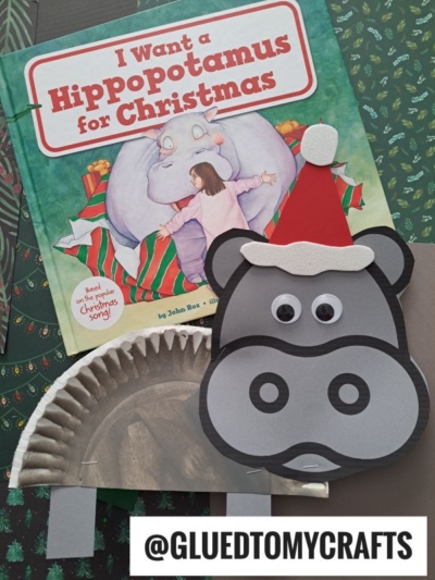 A children’s book titled “I Want a Hippopotamus for Christmas” is shown with a festive paper plate hippo craft, complete with a Santa hat. The handle @GLUEDTOMYCRAFTS appears at the bottom.