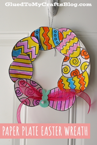 A colorful paper plate Easter egg wreath made from paper plates and decorated paper eggs, embellished with a pink ribbon and a small blue felt bunny, hangs on a white door. The text reads PAPER PLATE EASTER WREATH.