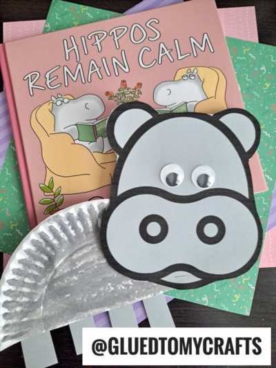 A children’s book titled Hippos Remain Calm sits on colored notebooks. In front, a cheerful paper plate hippo craft with googly eyes and gray paper features is displayed. A tag reads “@GLUEDTOMYCRAFTS.”.