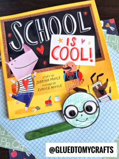 A cheerful children’s book titled School is Cool! is placed on patterned paper, alongside a handmade popsicle stick bookworm with a smiling face. The Instagram handle @GLUEDTOMYCRAFTS appears at the bottom.