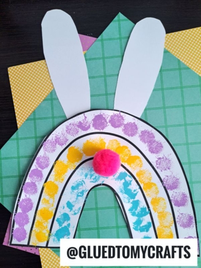 A colorful rainbow bunny paper craft features purple and yellow paint dots, two large white bunny ears, and a pink pom-pom nose, all set on patterned paper. Text reads @GLUEDTOMYCRAFTS.