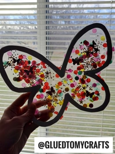 A hand holds up a butterfly suncatcher with colorful confetti and glitter inside, against a window with blinds. A label at the bottom reads @GLUEDTOMYCRAFTS.