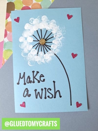 A blue card with a painted dandelion blowing in the wind, the words Make a wish, four red hearts, and the handle @GLUEDTOMYCRAFTS in a green box at the bottom—perfect inspiration for flower craft ideas.