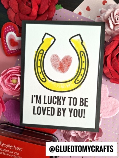 A handmade Fingerprint St. Patrick's Day card featuring a yellow horseshoe and red heart in the center. Below, it says IM LUCKY TO BE LOVED BY YOU! The card is surrounded by red and pink paper flowers and craft supplies.