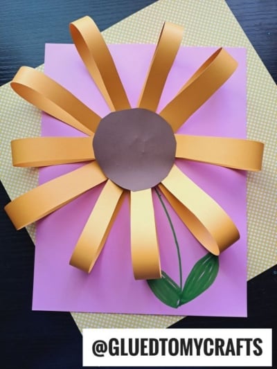 A handmade flower craft, perfect for flower kids crafts, features yellow paper loop petals, a brown paper center, and a green drawn stem on a pink sheet with a yellow-patterned background. Text reads @GLUEDTOMYCRAFTS.