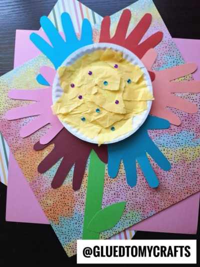 A colorful paper flower kids craft made with layered, cut-out handprints for petals, a green stem, and a center of yellow tissue paper on a paper plate, decorated with small gems. A watermark reads @GLUEDTOMYCRAFTS.