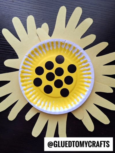 A sunflower craft made from a yellow paper plate with black dots in the center, layered on top of yellow paper handprints shaped like petals. Perfect for flower kids crafts, @GLUEDTOMYCRAFTS is written at the bottom.