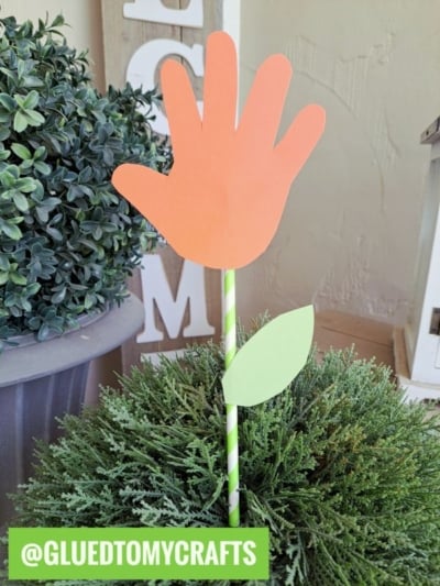 A fun flower kids crafts project: an orange handprint cutout forms the flower on a striped straw stem, complete with a green paper leaf. Displayed in a potted plant and labeled @GLUEDTOMYCRAFTS for creative flower craft ideas!.
