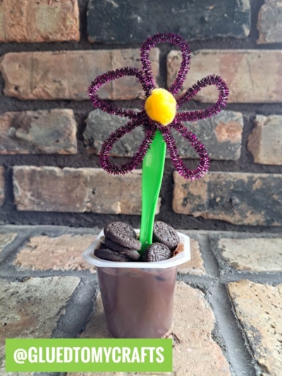 A fun flower kids craft: A purple pipe cleaner and yellow pom-pom flower is attached to a green spoon, set in a chocolate pudding cup with mini cookies, against a brick wall. @GLUEDTOMYCRAFTS is labeled at the bottom.